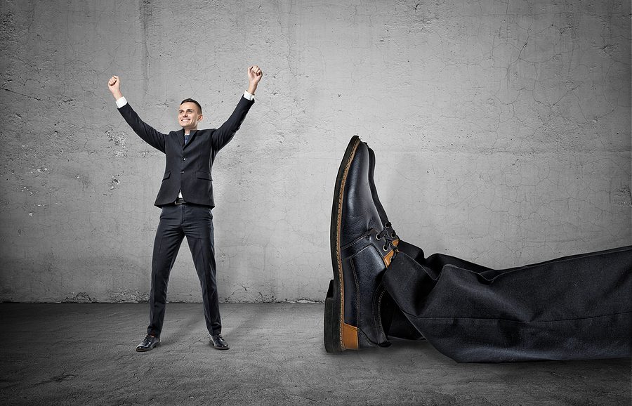 A small businessman standing with his arms up and smiling happily near a giant leg in trousers and shoe lying near him, on the concrete gray background. Winning contest. Best in field. David and Goliath. Small company wins over big one. M-Files